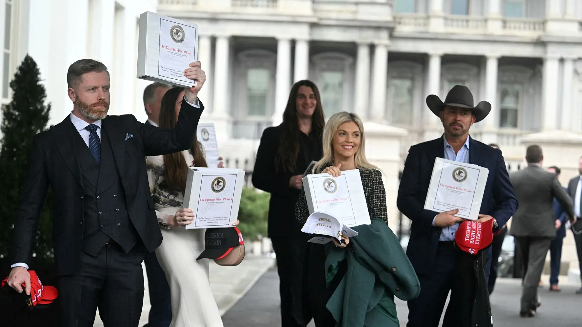 مجموعة من الشخصيات الإعلامية المؤيدة لترامب تحمل مجلدات مكتوب عليها "ملفات إبشتاين: المرحلة 1" خارج البيت الأبيض، وسط أجواء من التوتر والترقب.