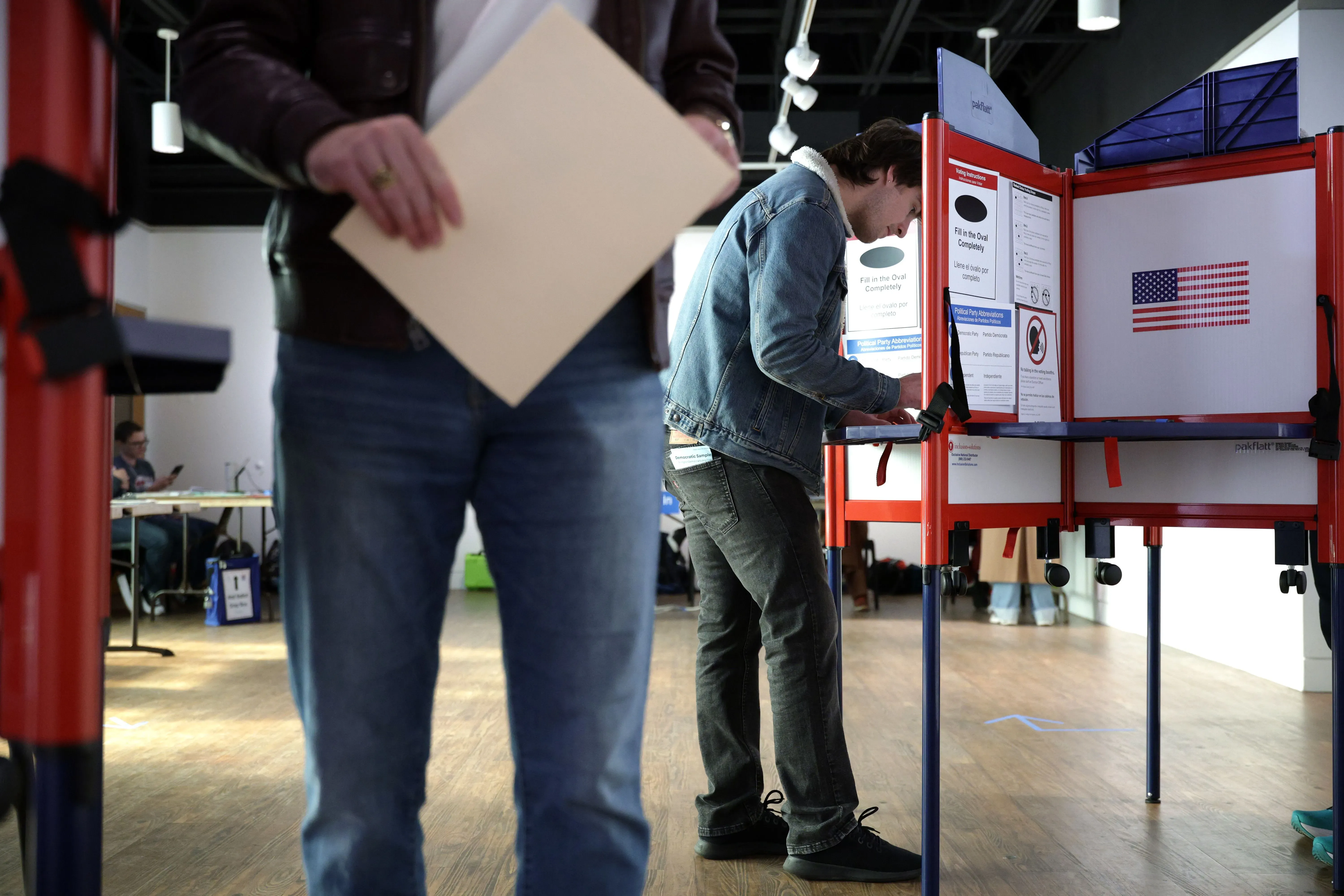 ناخب يقف أمام آلة التصويت في مركز اقتراع، حاملًا ورقة، بينما يظهر في الخلف أشخاص آخرون. تعكس الصورة أهمية التصويت في سياق النقاش حول قانون إنقاذ أمريكا.