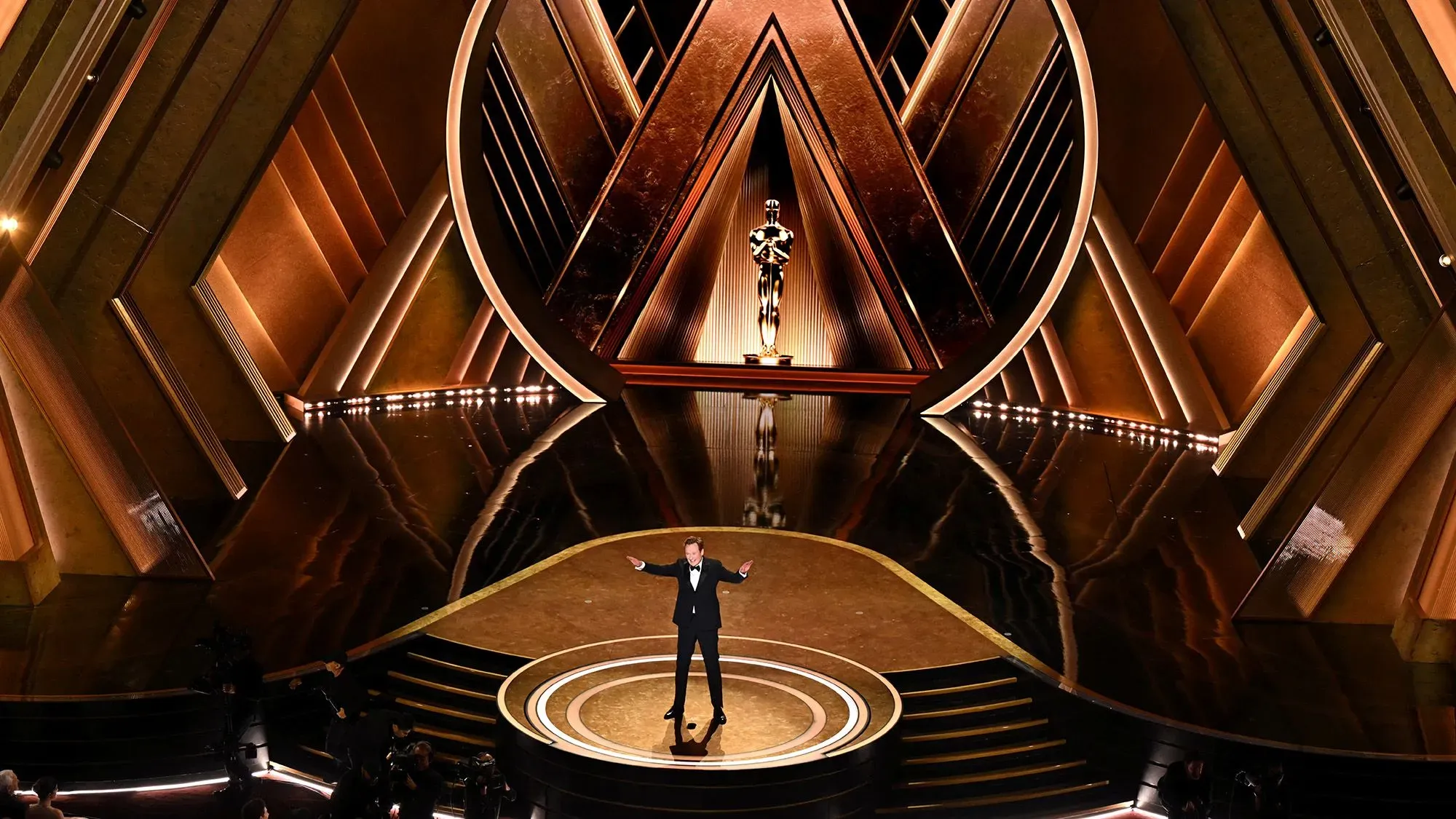 مقدم حفل الأوسكار يقف على المسرح أمام تمثال الأوسكار، مع تصميم داخلي أنيق يبرز الأضواء والديكورات الذهبية.