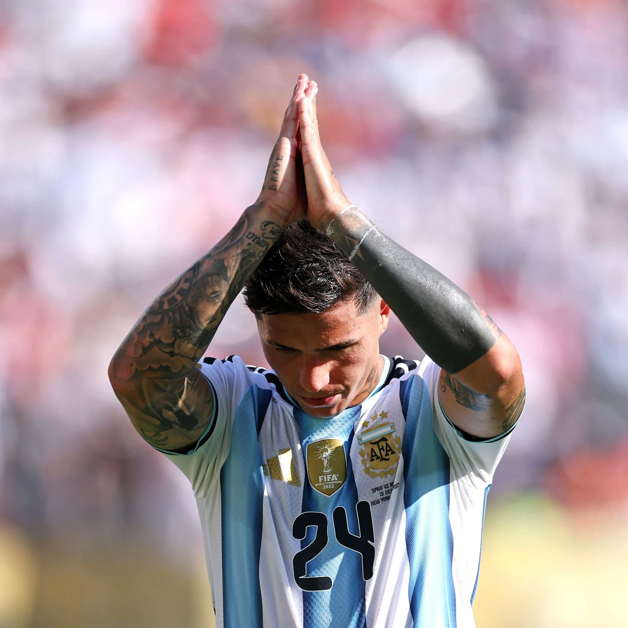 لاعب كرة قدم أرجنتيني يرتدي قميص منتخب بلاده يرفع يديه متشابكتين في تحية أو شكر خلال مباراة، مع خلفية ضبابية للجمهور.