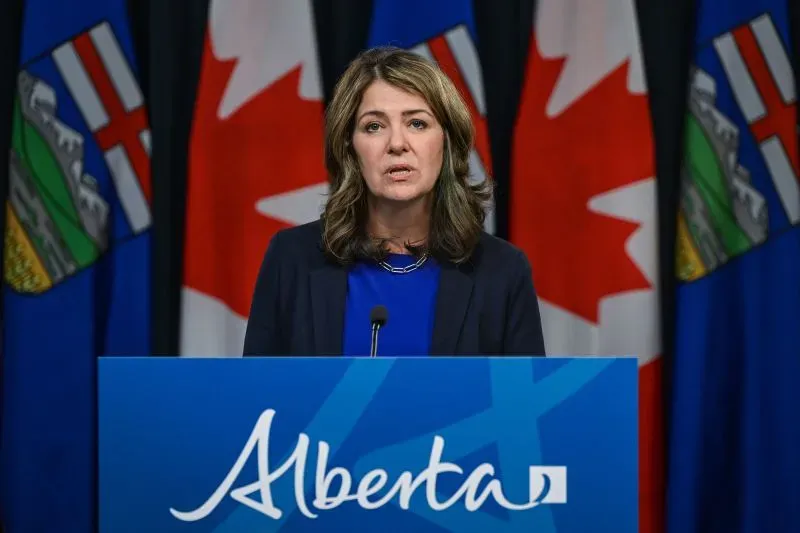 امرأة تتحدث في مؤتمر صحفي، أمام لافتة تحمل اسم "ألبرتا"، وخلفها أعلام كندية وألبرتا، تعبر عن قضايا الاستقلال.