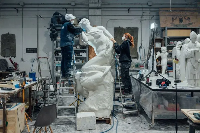 عمال في ورشة عمل يقومون بترميم منحوتة من الجبس لمايكل أنجلو، مع وجود تماثيل أخرى في الخلفية، في سياق معرض فني.