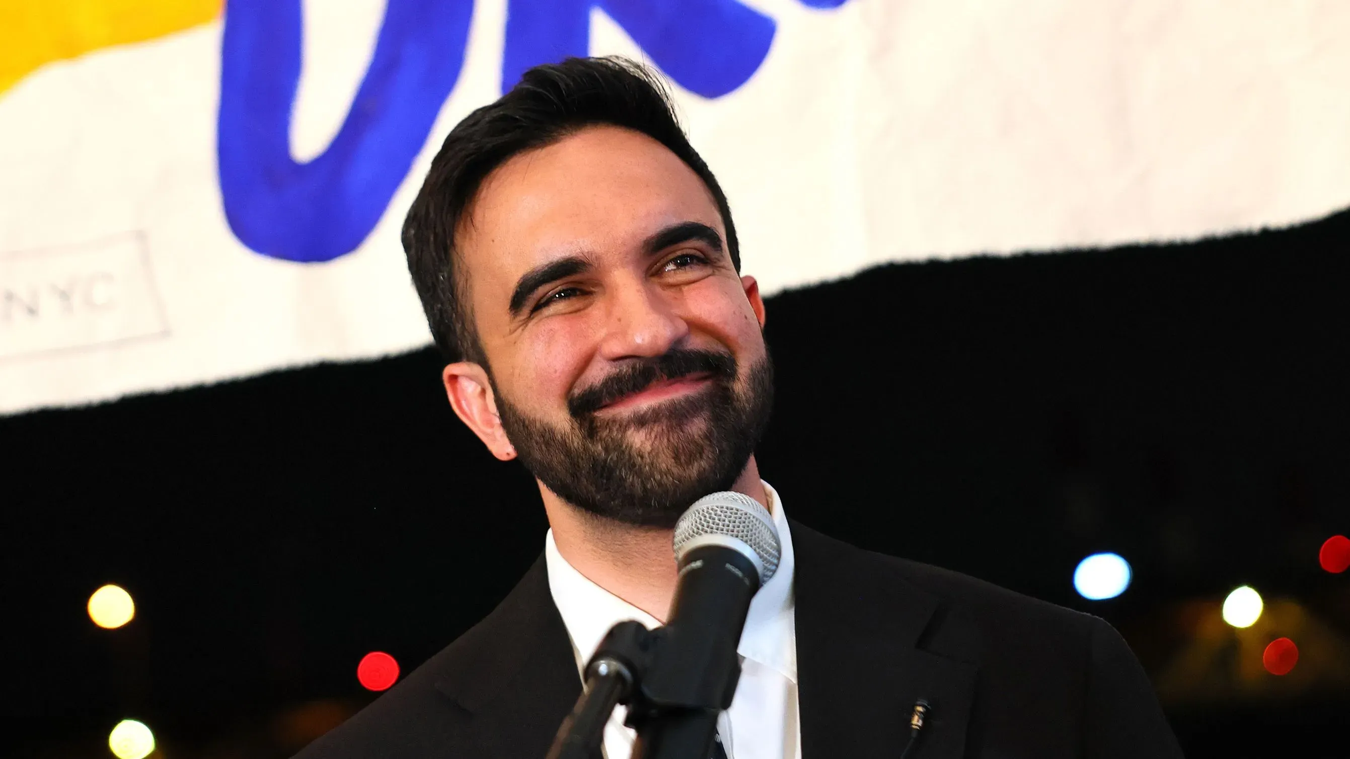 زهران ممداني، المرشح الديمقراطي لعمودية نيويورك، مبتسم أثناء حديثه في ليلة الانتخابات، مع وجود لافتة خلفه.