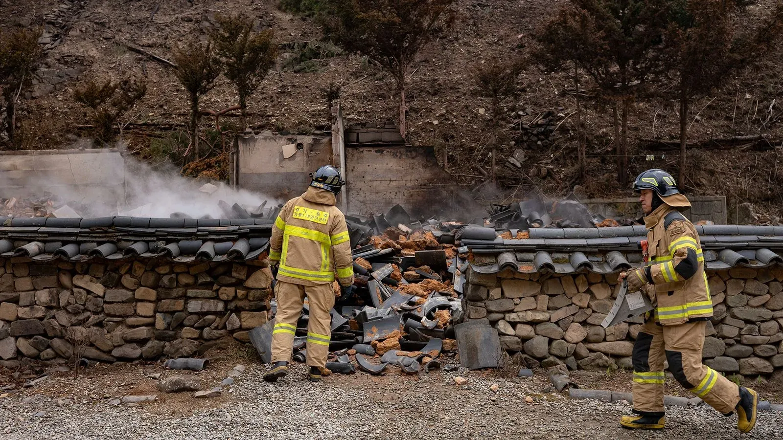 رجال إطفاء يتفقدون حطام مبنى دمرته حرائق الغابات في كوريا الجنوبية، مع تصاعد الدخان في الخلفية.