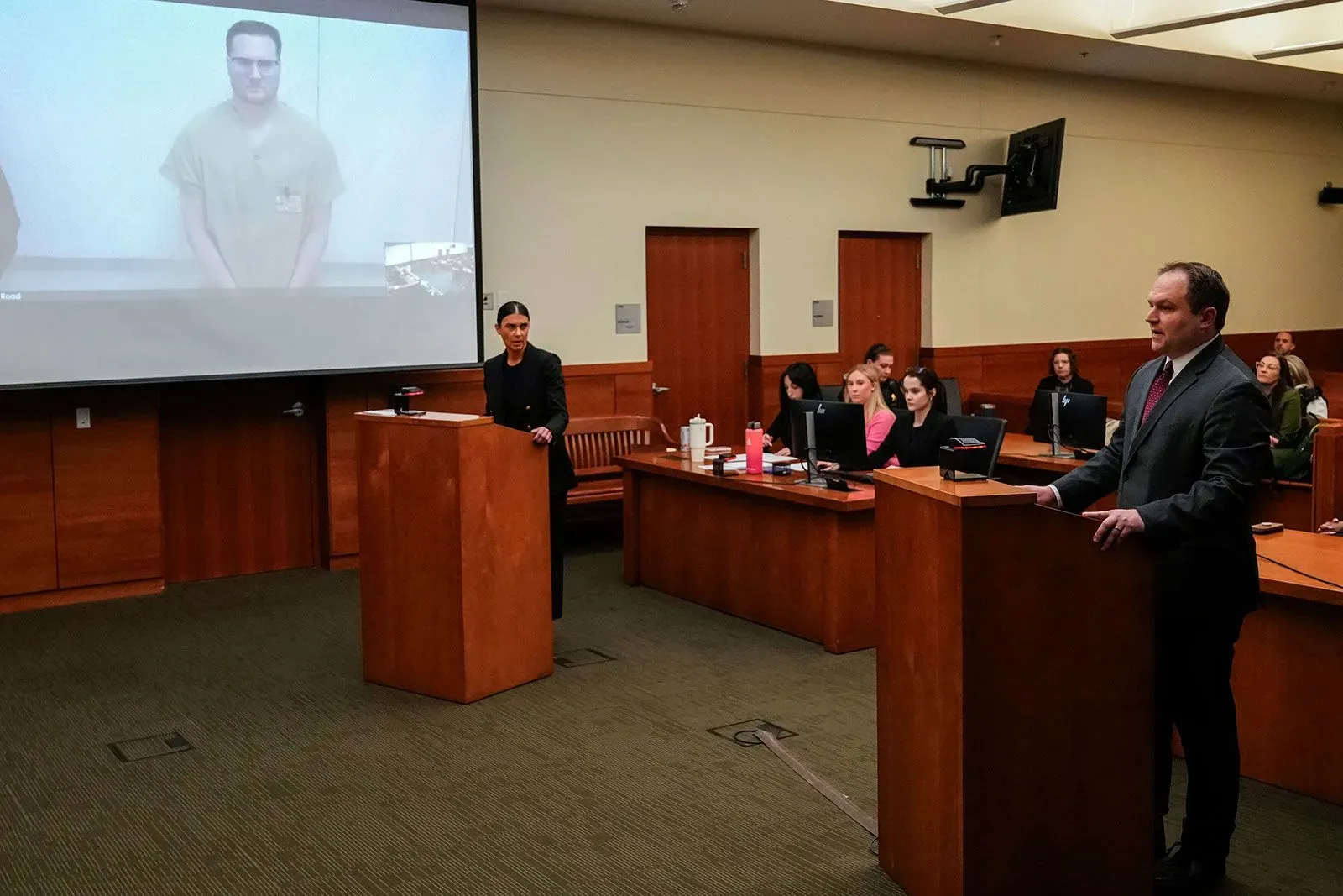 جلسة محكمة تُظهر المتهم مايكل ماكي عبر الفيديو، مع محاميه أمام القاضي، بينما يتواجد آخرون في قاعة المحكمة.