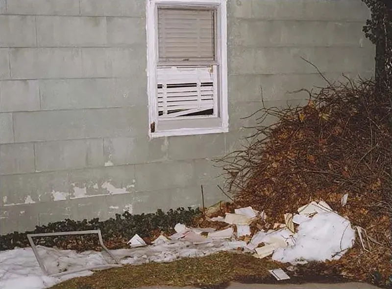 صورة توضح نافذة مكسورة لمنزل إدنا شوبرت، مع أكوام من الأوراق والثلوج المتراكمة بجانبها، مما يعكس مشهد جريمة القتل الغامضة.