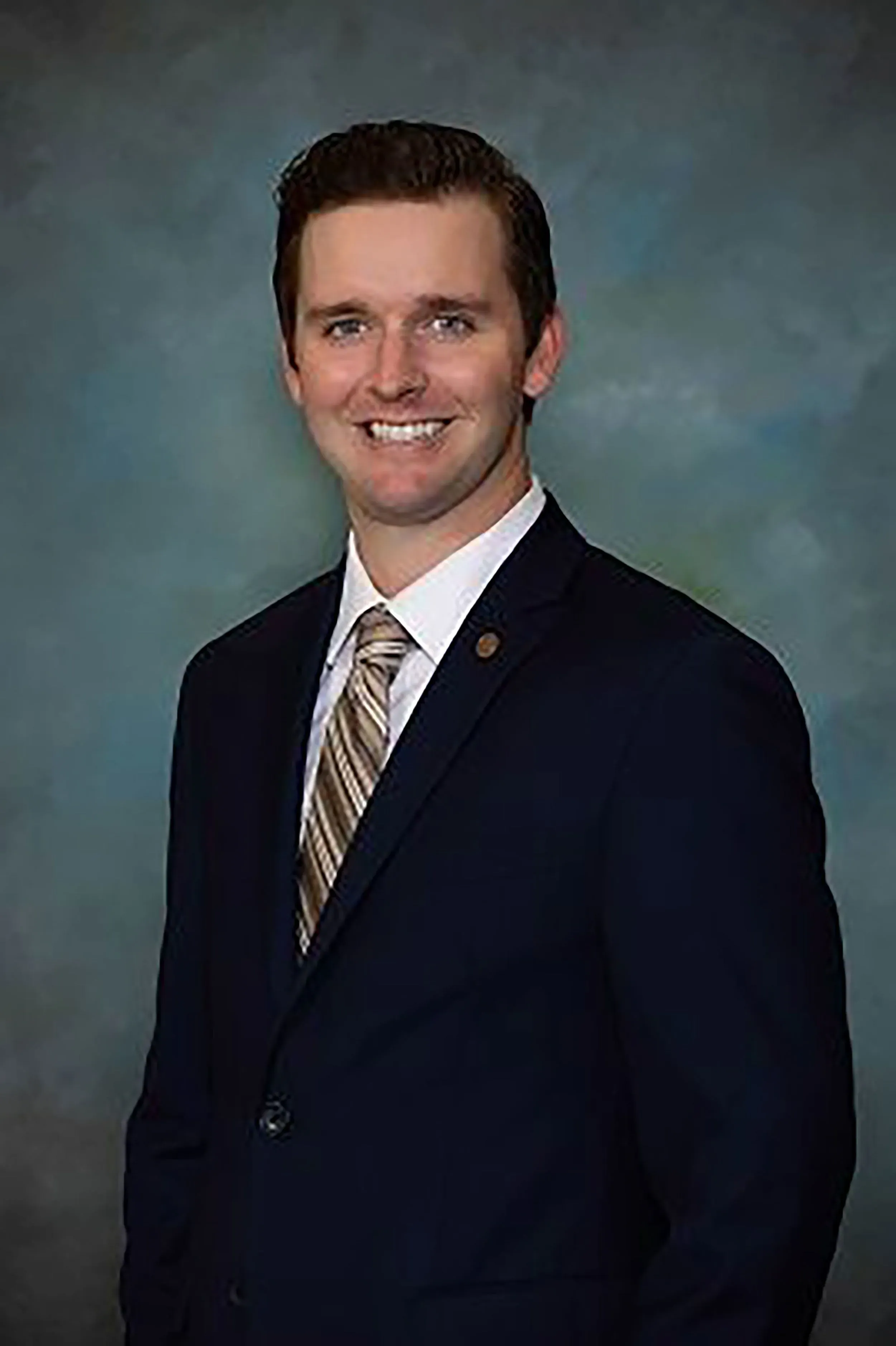 صورة لرجل مبتسم يرتدي بدلة رسمية، يظهر في خلفية رمادية، في سياق حادث اعتداء على عضو مجلس مدينة دانفيل.