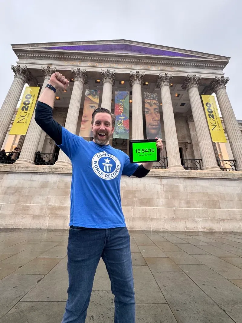 رجل مبتسم يرتدي قميص موسوعة غينيس للأرقام القياسية، يقف أمام متحف معروف في لندن حاملًا جهاز توقيت، بعد زيارته لـ 42 متحفًا في أقل من 12 ساعة.