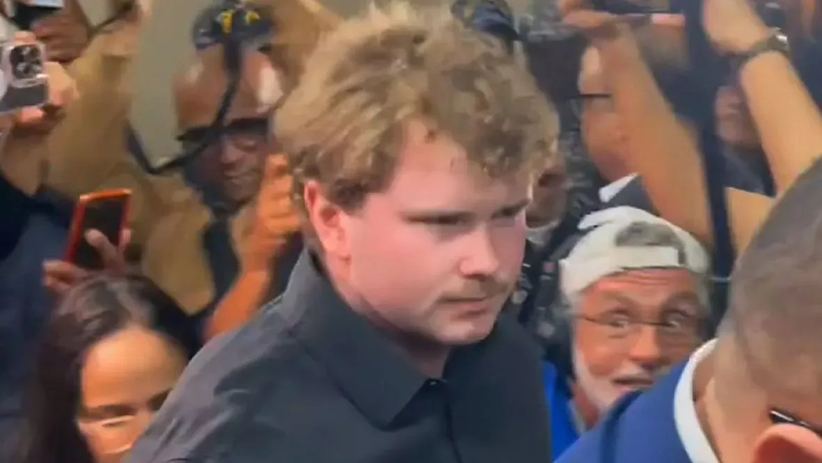 شخص شاب ذو شعر أشقر ومجعد، يرتدي قميصًا أسود، يظهر وسط حشد من الناس مع هواتف محمولة، بعد جلسة استماع في المحكمة حول قضية طالبة مفقودة.