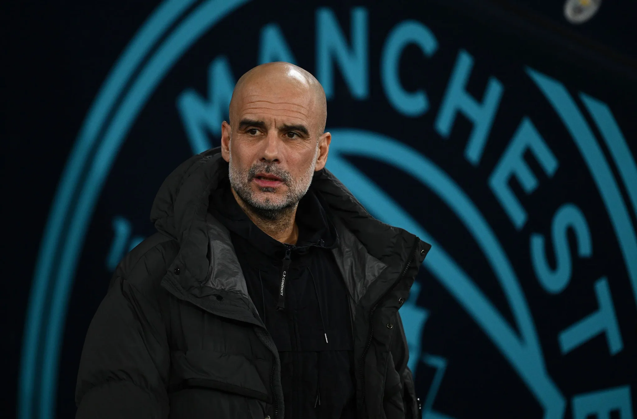 بيب غوارديولا مدرب مانشستر سيتي أمام شعار النادي، في ظل التحقيقات المالية التي تهدد ألقاب الفريق الإنجليزي.