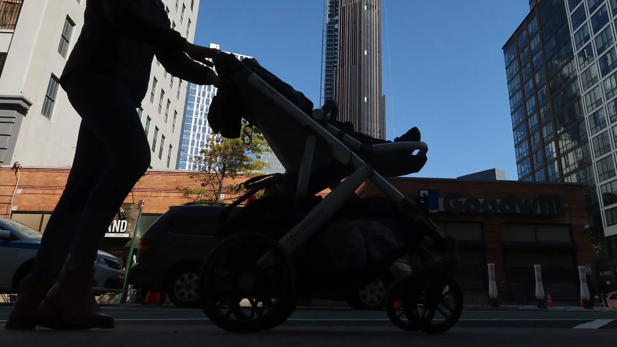 امرأة تدفع عربة أطفال في مدينة، مع خلفية لمبانٍ عالية، تعكس تحديات العمل في رعاية الأطفال.