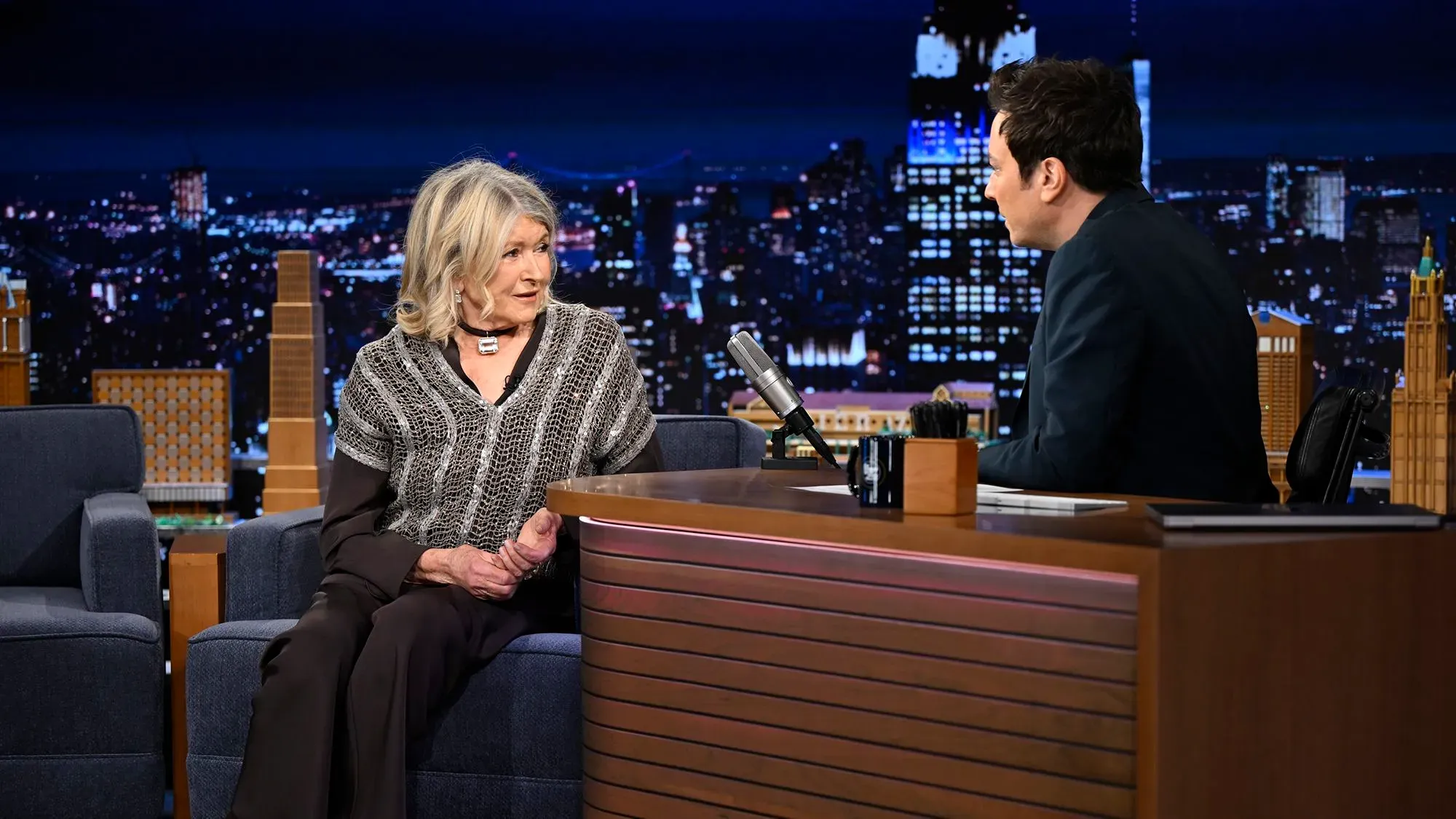 مارثا ستيوارت تتحدث مع جيمي فالون في برنامجه "ذا تونايت شو"، مع خلفية مميزة لمدينة نيويورك ليلاً، تعكس أجواء العرض.