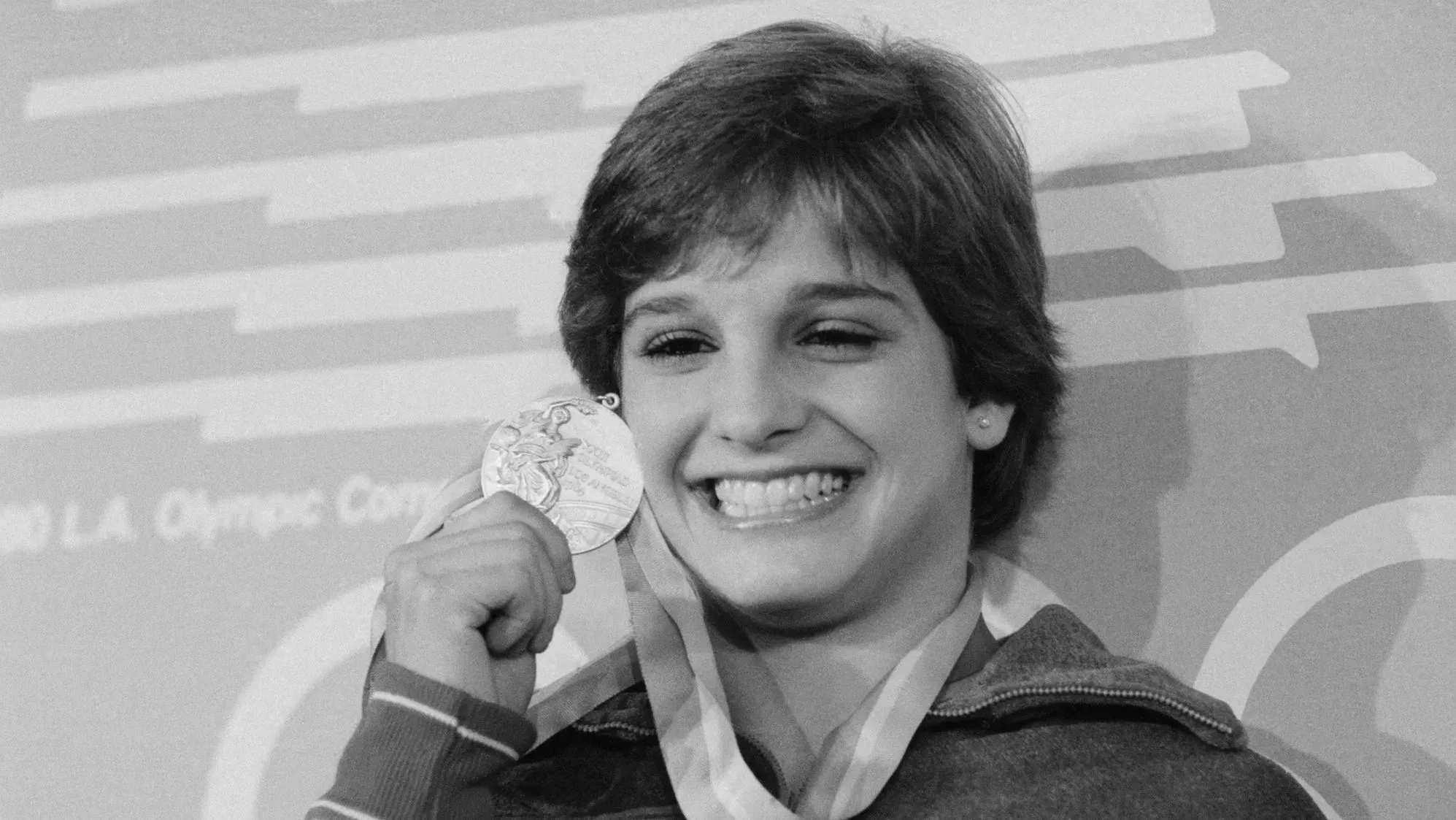 ماري لو ريتون، أيقونة الجمباز الأمريكية، تبتسم بفخر وهي تحمل ميداليتها الأولمبية، مما يعكس إنجازاتها الرياضية التاريخية.
