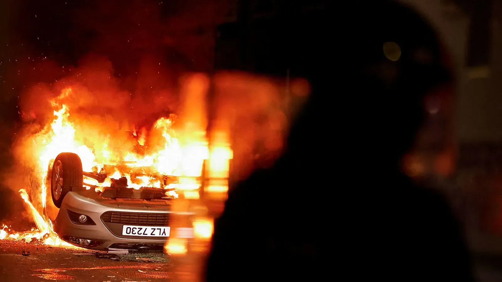 سيارة مشتعلة مقلوبة في بلدة باليمينا، مع وجود ضابط شرطة في الخلفية، في مشهد من أعمال الشغب العنيفة التي تلت احتجاجًا على اعتداء جنسي مزعوم.