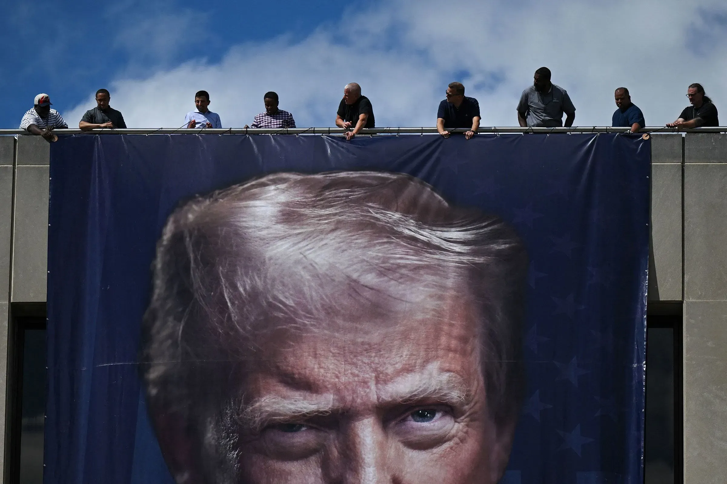 لافتة كبيرة تحمل صورة الرئيس ترامب، مع مجموعة من الأشخاص يعملون على تثبيتها على حافة مبنى فيدرالي.