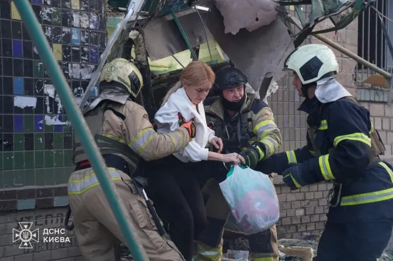 رجال إطفاء يساعدون امرأة على الخروج من مبنى متضرر بعد هجوم بطائرات مسيرة في كييف، مما يعكس آثار الهجمات الروسية على المدنيين.