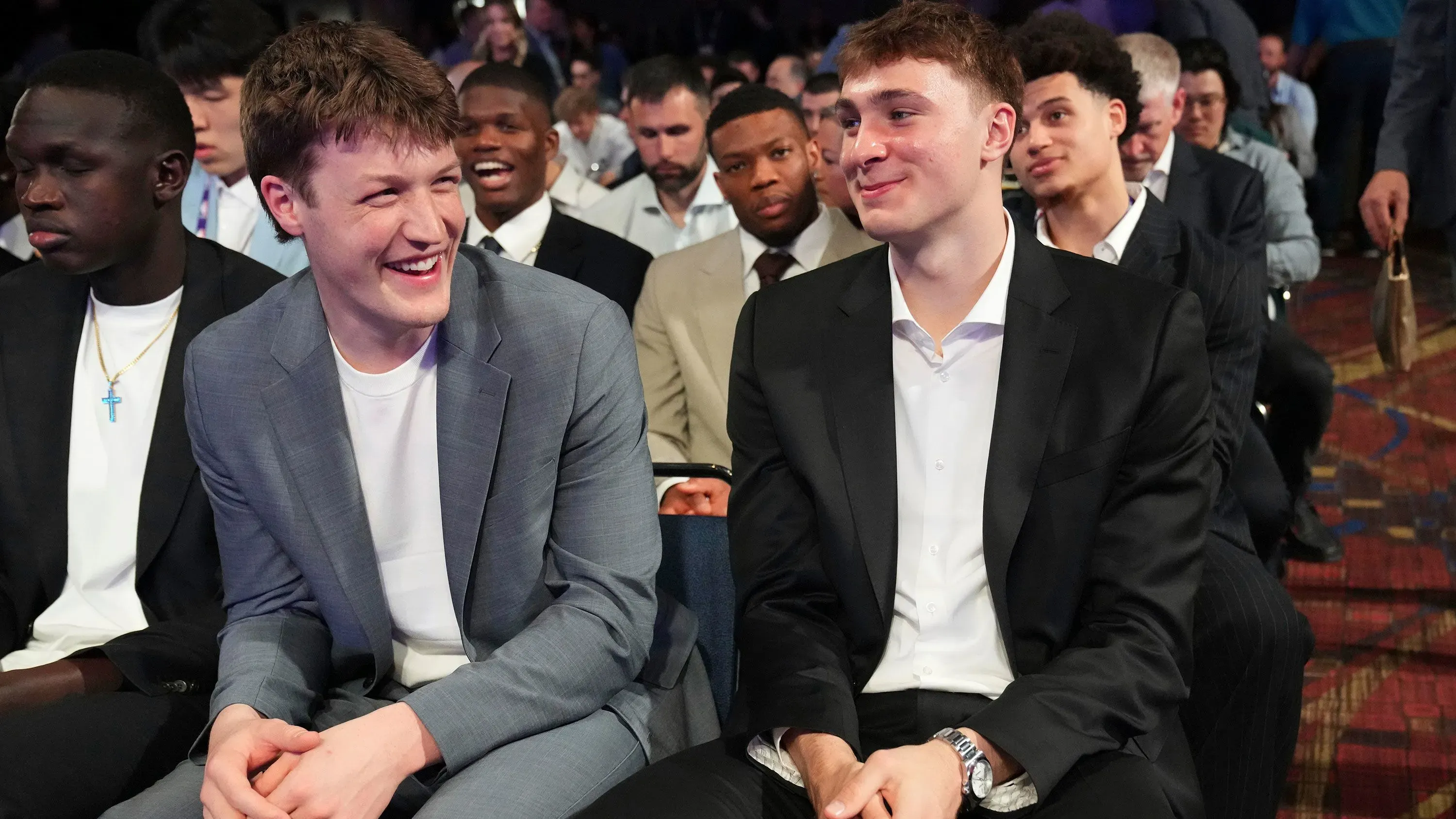 شباب مبتسمون يرتدون ملابس رسمية، يجلسون معًا في حدث رياضي، مع تواجد مشجعين آخرين في الخلفية، يعكسون أجواء الترقب في مسودة الدوري الأمريكي للمحترفين.