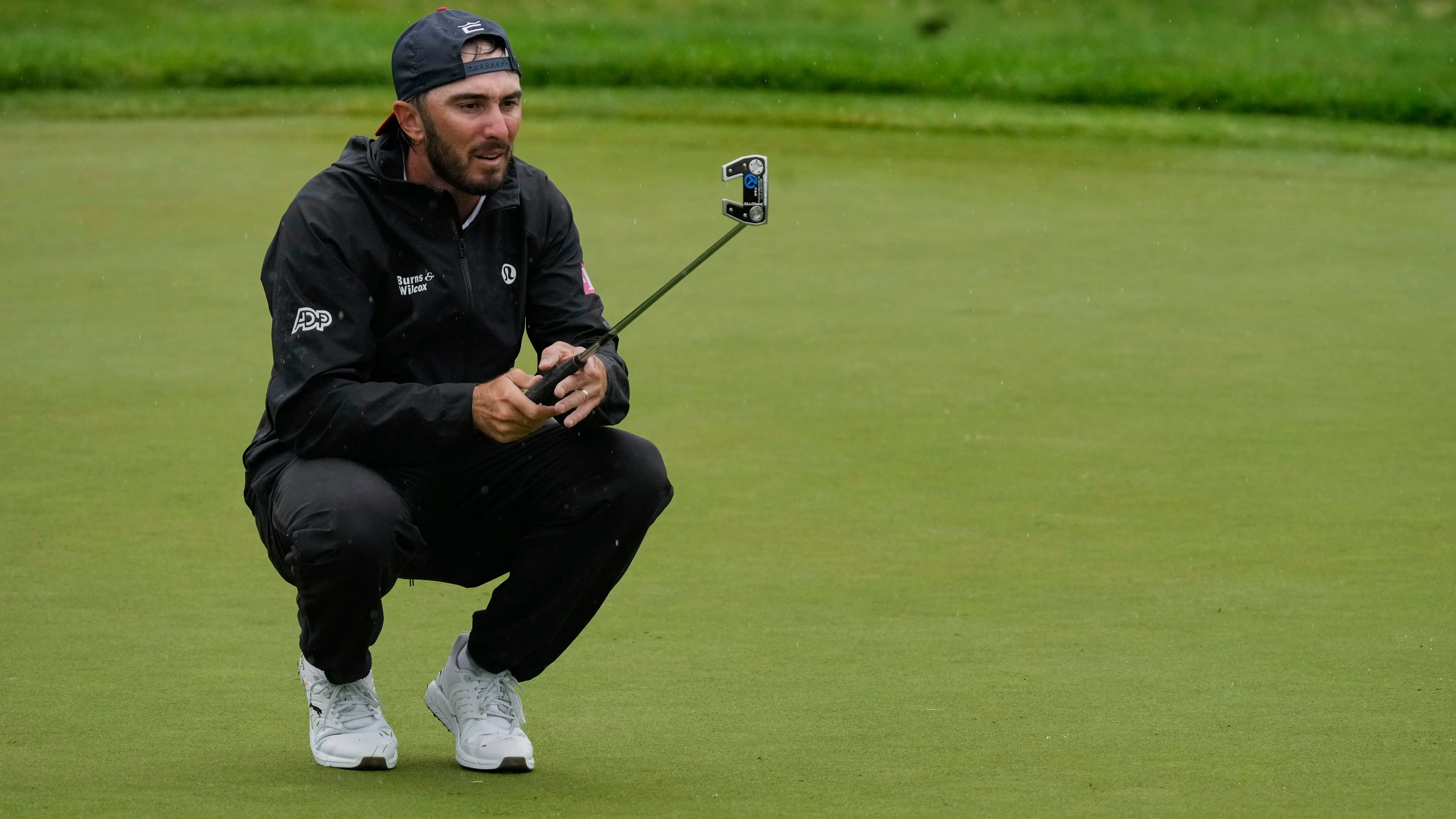 ماكس هوما يتأهب لتسديد كرة الغولف على ملعب كينسيل، مرتديًا ملابس رياضية سوداء، مع التركيز على وضعه في التصفيات لبطولة الولايات المتحدة المفتوحة.