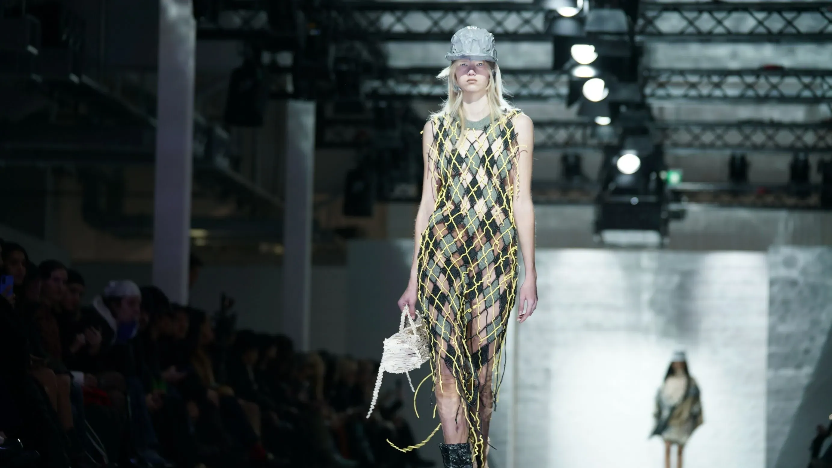 عارضة أزياء ترتدي فستانًا شبكيًا مبتكرًا من اللاتكس، بينما تحمل حقيبة من تصميم كاسيا كوتشارسكا خلال عرض أزياء في برلين.