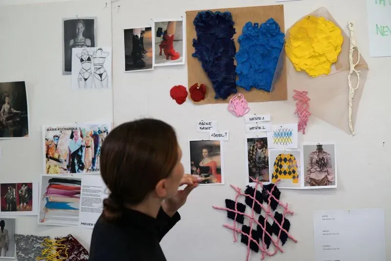 امرأة تفكر أمام لوحة تحتوي على تصاميم أزياء ملونة وصور، تعكس إبداع المصممة كاسيا كوتشارسكا في استخدام اللاتكس.