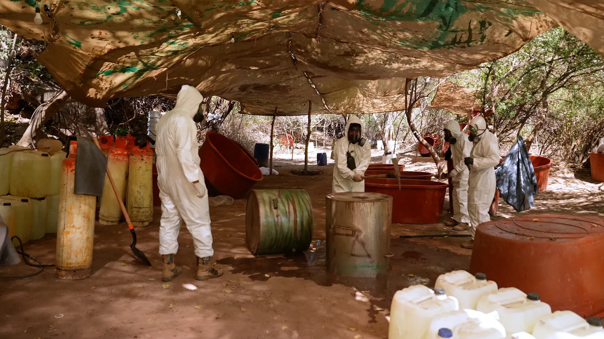 عمال يرتدون بدلات واقية يعملون في موقع سري لإنتاج الفنتانيل، محاطين بأواني ومواد كيميائية تحت غطاء في منطقة نائية بالمكسيك.