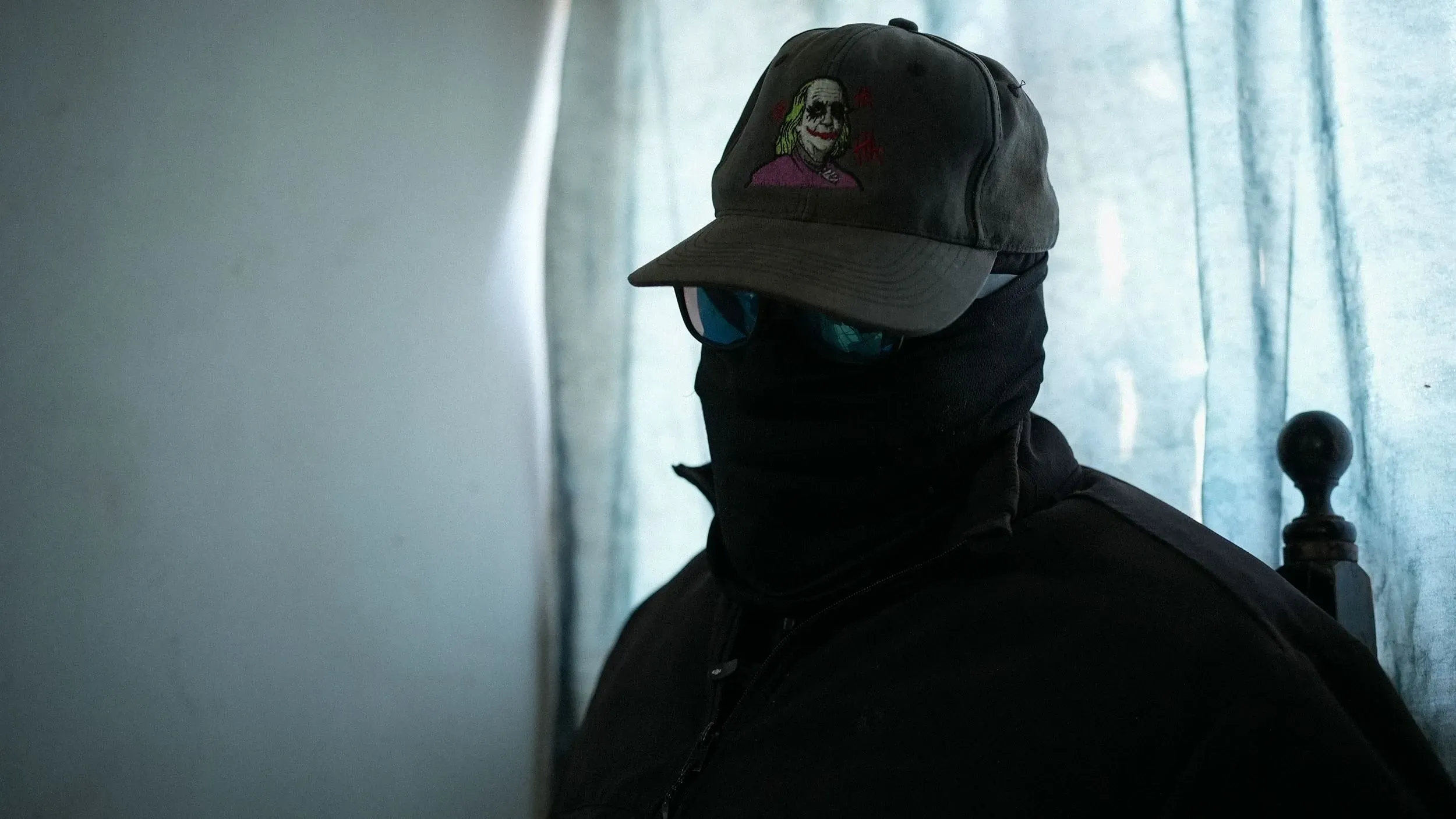 رجل ملثم يجلس في غرفة منخفضة الإضاءة، يرتدي قبعة بيسبول ونظارات شمسية، محاطًا بأجهزة اتصال وبندقية، ويعبر عن تحديات الكارتل في إنتاج الفنتانيل.