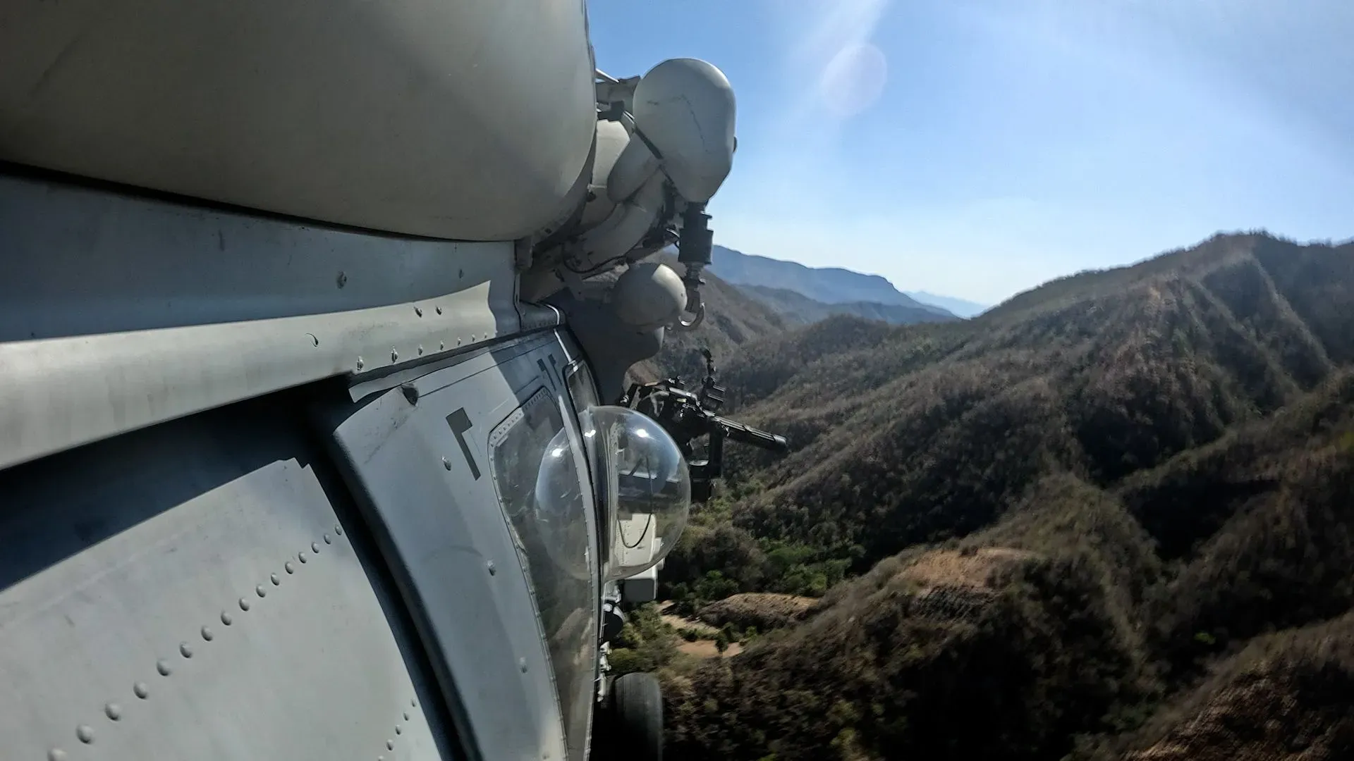 صورة تظهر طائرة هليكوبتر عسكرية تحلق فوق جبال في المكسيك، مع جندي مسلح يستعد لمراقبة الأنشطة الأرضية.