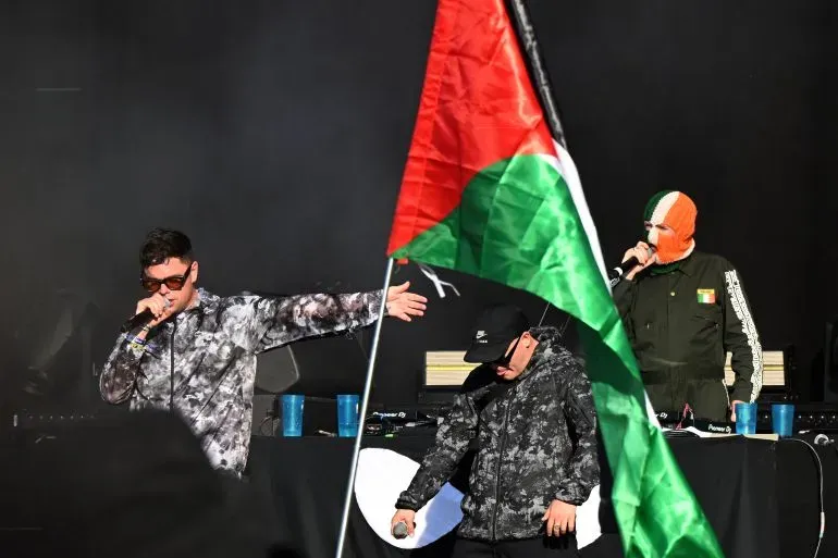 يظهر في الصورة أعضاء فرقة "كنيكاب" يؤدون عرضًا موسيقيًا، بينما يرفع أحدهم علم فلسطين. تعكس الصورة دعمهم للقضية الفلسطينية وسط جدل حول التهم الموجهة لأحدهم.