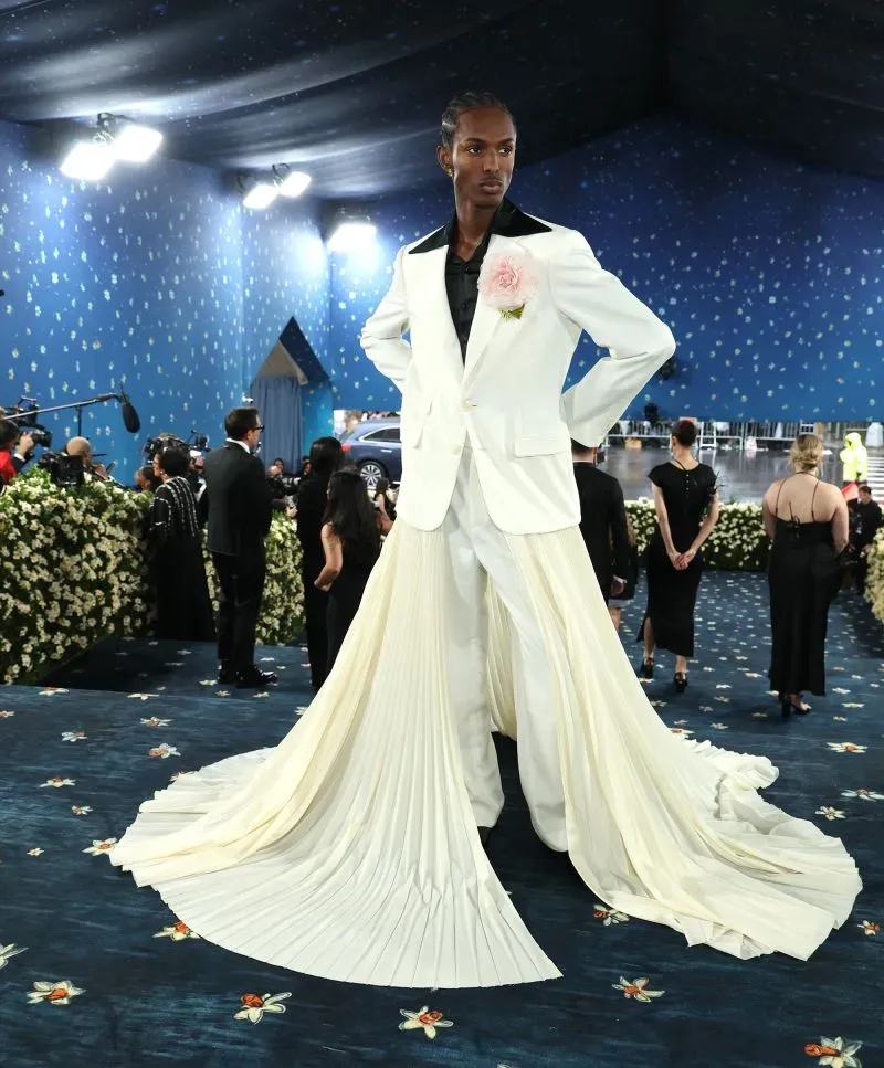 عارض أزياء يرتدي بدلة بيضاء مع تنورة طويلة من القماش المجعد، يقف في حفل الميت غالا، مع خلفية زرقاء وزهور بيضاء.