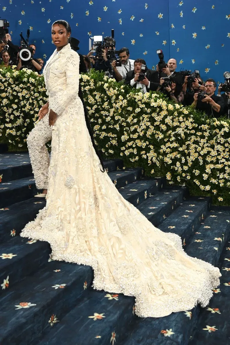 نجم يرتدي فستانًا أبيض مزينًا بالتفاصيل، يتجه نحو السلالم في حفل الميت غالا، محاطًا بكاميرات المصورين وزهور بيضاء.