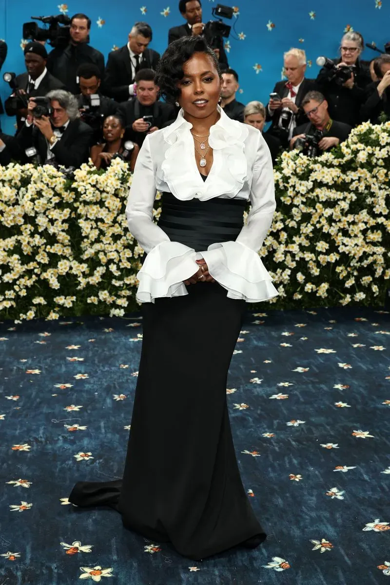 نجمة ترتدي فستاناً أنيقاً باللون الأسود مع تفاصيل بيضاء، تتوسط حفل الميت غالا في متحف متروبوليتان، محاطة بزهور بيضاء وصحافة.