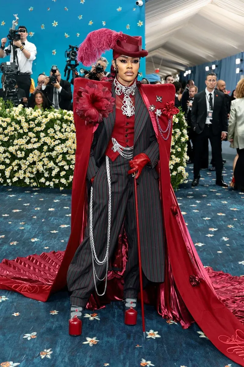 إطلالة جريئة لنجمة ترتدي بدلة مخططة مع عباءة حمراء وقبعة مزينة، في حفل ميت غالا بمتحف متروبوليتان، محاطة بكاميرات المصورين.