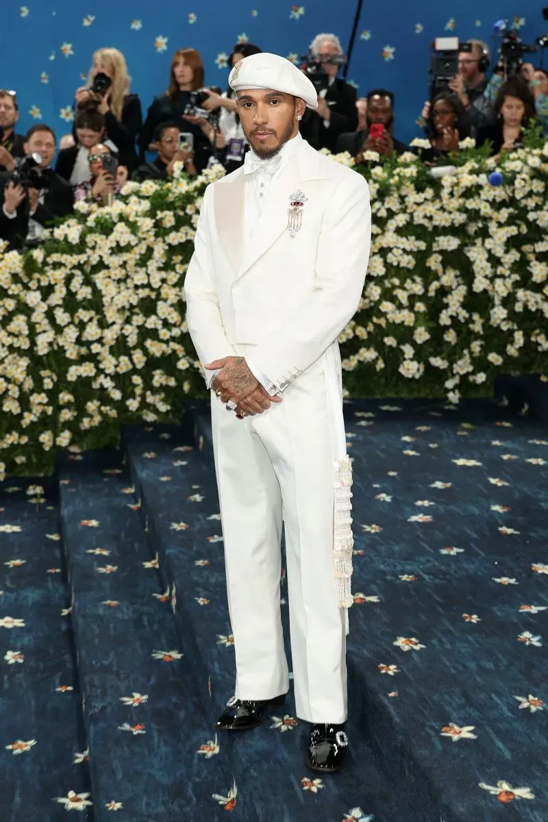 لويس هاميلتون يرتدي بدلة بيضاء أنيقة في حفل الميت غالا، محاطًا بديكور زهور، وسط عدسات الكاميرات.