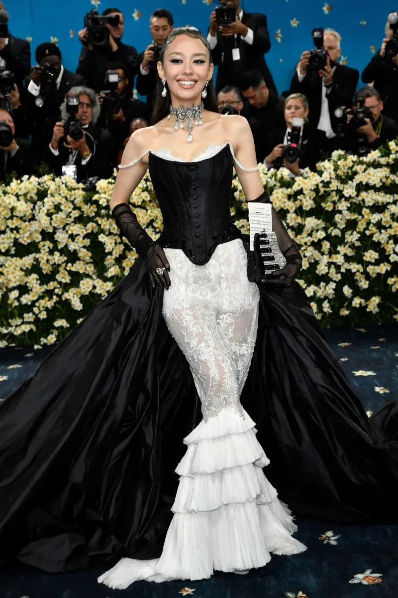 نجمة ترتدي فستانًا أسود وأبيض مذهل في حفل الميت غالا، محاطة بزهور بيضاء وعدسات الكاميرا، تعكس أناقة الحدث الثقافي.