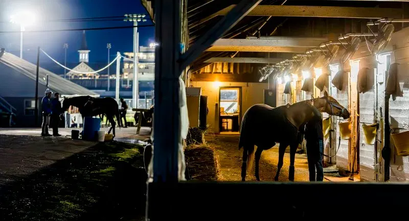 إسطبلات مضمار تشرشل داونز مضاءة ليلاً، مع وجود خيول وفريق عمل يستعدون لسباق كنتاكي ديربي الشهير.