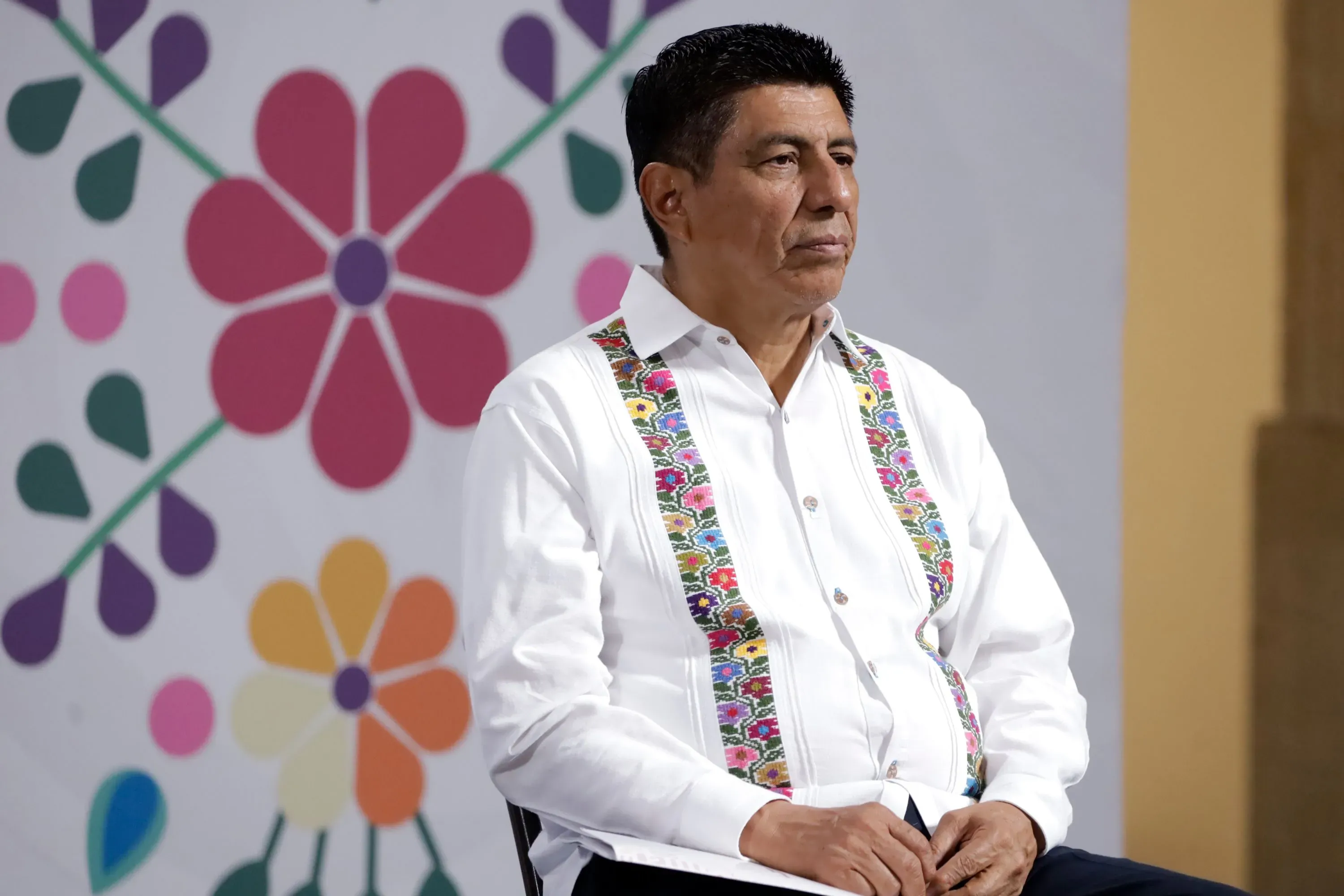 حاكم ولاية أواكساكا، سالومون خارا كروز، يجلس في مؤتمر صحفي، مع خلفية مزخرفة بالألوان، يتحدث عن قضية استيلاء ثقافي من أديداس.