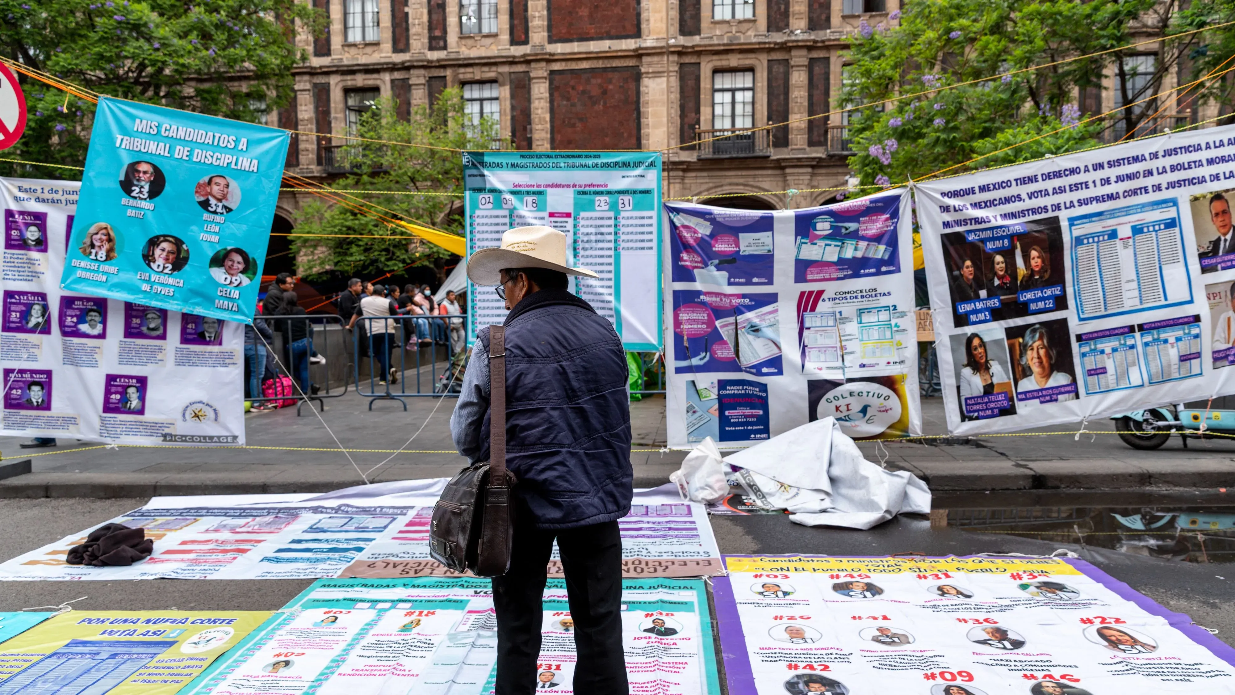 رجل يرتدي قبعة يقف أمام ملصقات انتخابية متعددة تتعلق بالانتخابات القضائية في المكسيك، مع تفاصيل عن المرشحين والمعلومات المهمة.