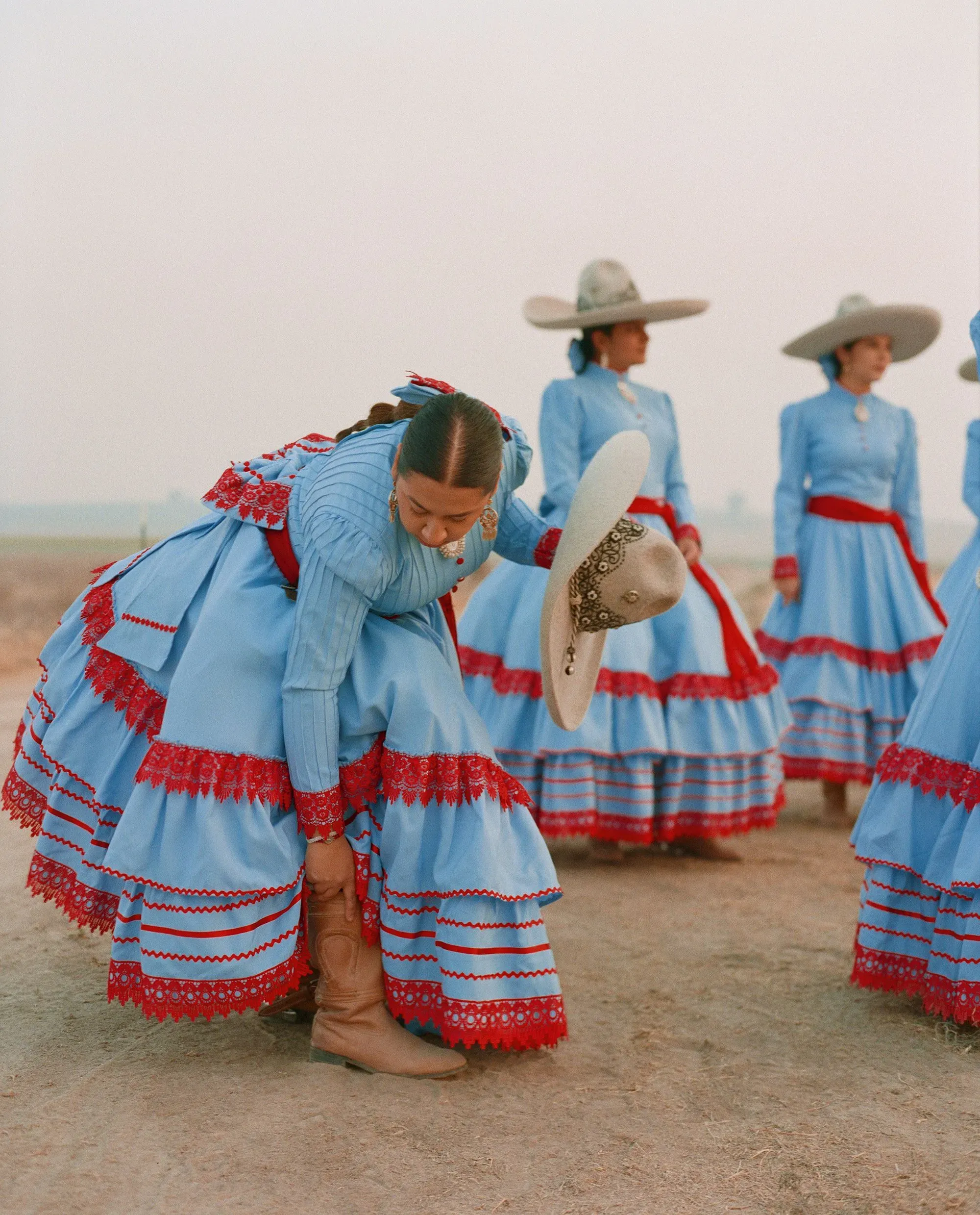 امرأة ترتدي فستانًا أزرق مزينًا بلون أحمر، تنحني لربط حذائها بينما تتجمع نساء أخريات في خلفية الصورة، استعدادًا لمسابقة إسكاراموزا.