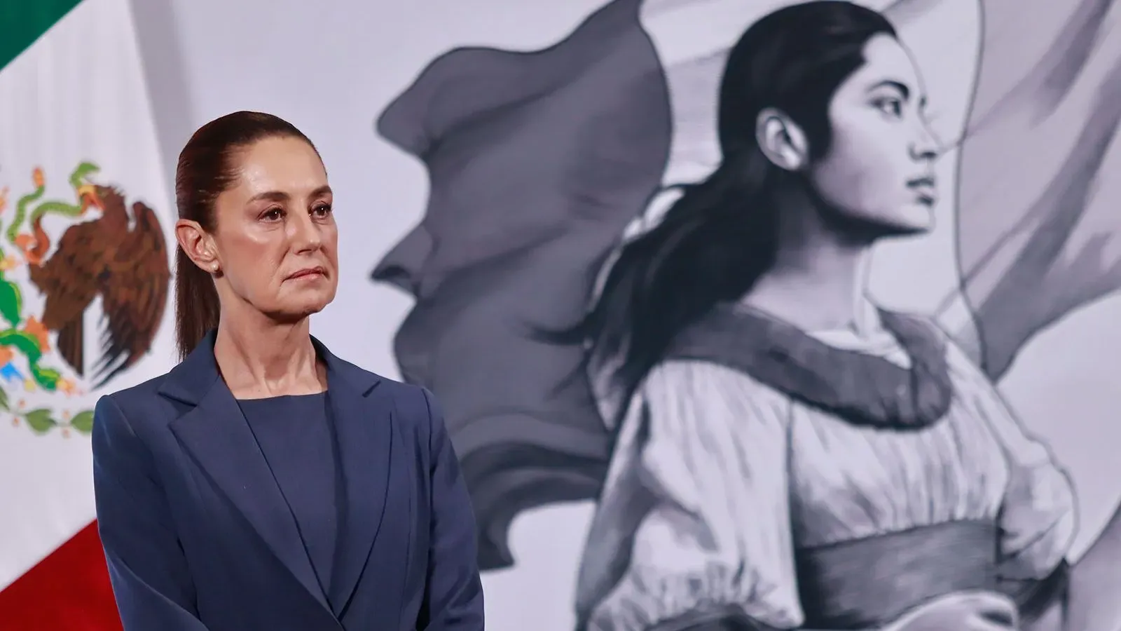 الرئيسة المكسيكية كلاوديا شينباوم تقف أمام لوحة تجسد امرأة تعبر عن القوة، مع العلم المكسيكي في الخلفية، خلال مؤتمر صحفي حول إعلانات الهجرة.