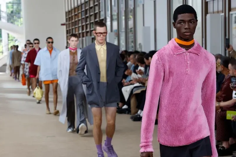 عرض أزياء رجالية في ميلانو، حيث يسير عارضون بملابس ملونة متنوعة، مع جمهور متنوع في الخلفية.