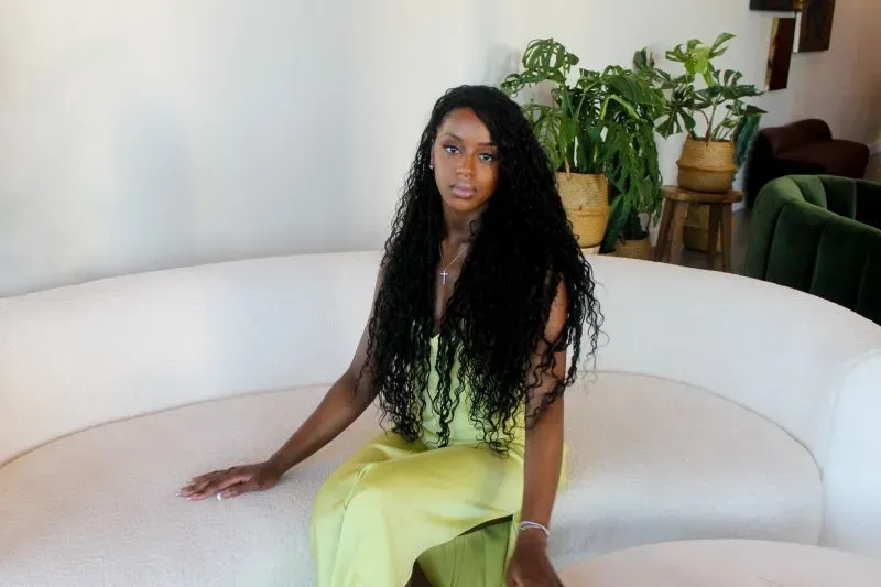 امرأة ذات شعر طويل ومجعد ترتدي فستانًا أصفر، تجلس على أريكة بيضاء في بيئة داخلية مريحة، محاطة بنباتات خضراء.