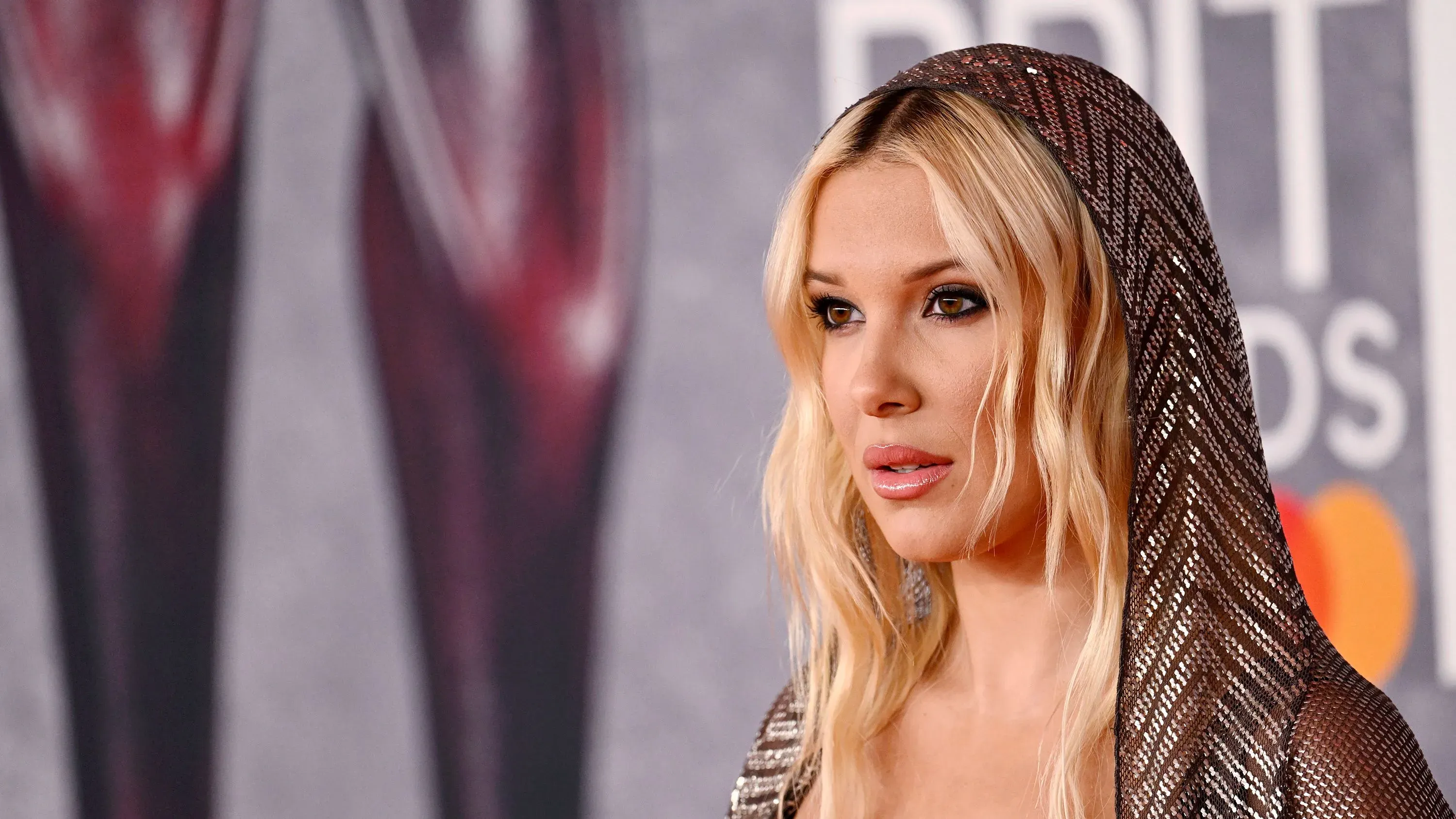 ميلي بوبي براون في حدث رسمي، ترتدي غطاء رأس لامع، وتظهر تعبيرًا مفاجئًا أثناء مناقشتها اسمها الأوسط "بوني".