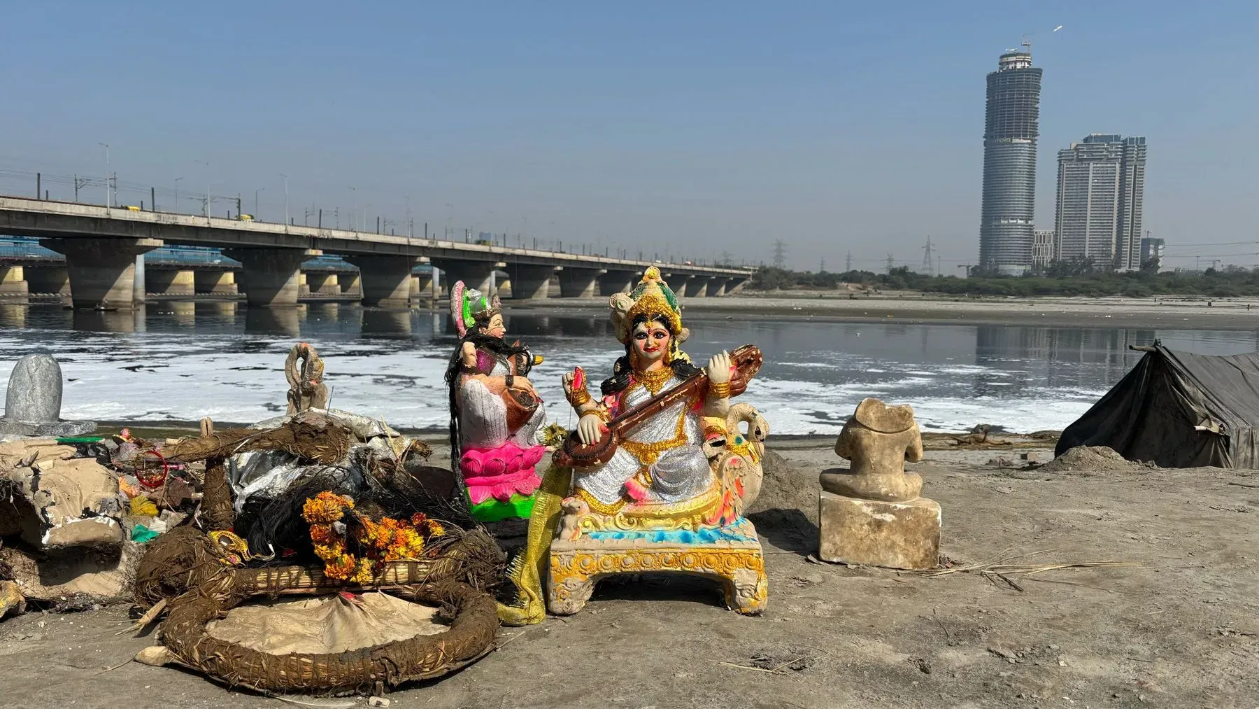 تمثال لآلهة هندوسية محاط بالنفايات على ضفاف نهر يامونا الملوث، مع خلفية تظهر جسرًا ومبانٍ حديثة في دلهي.