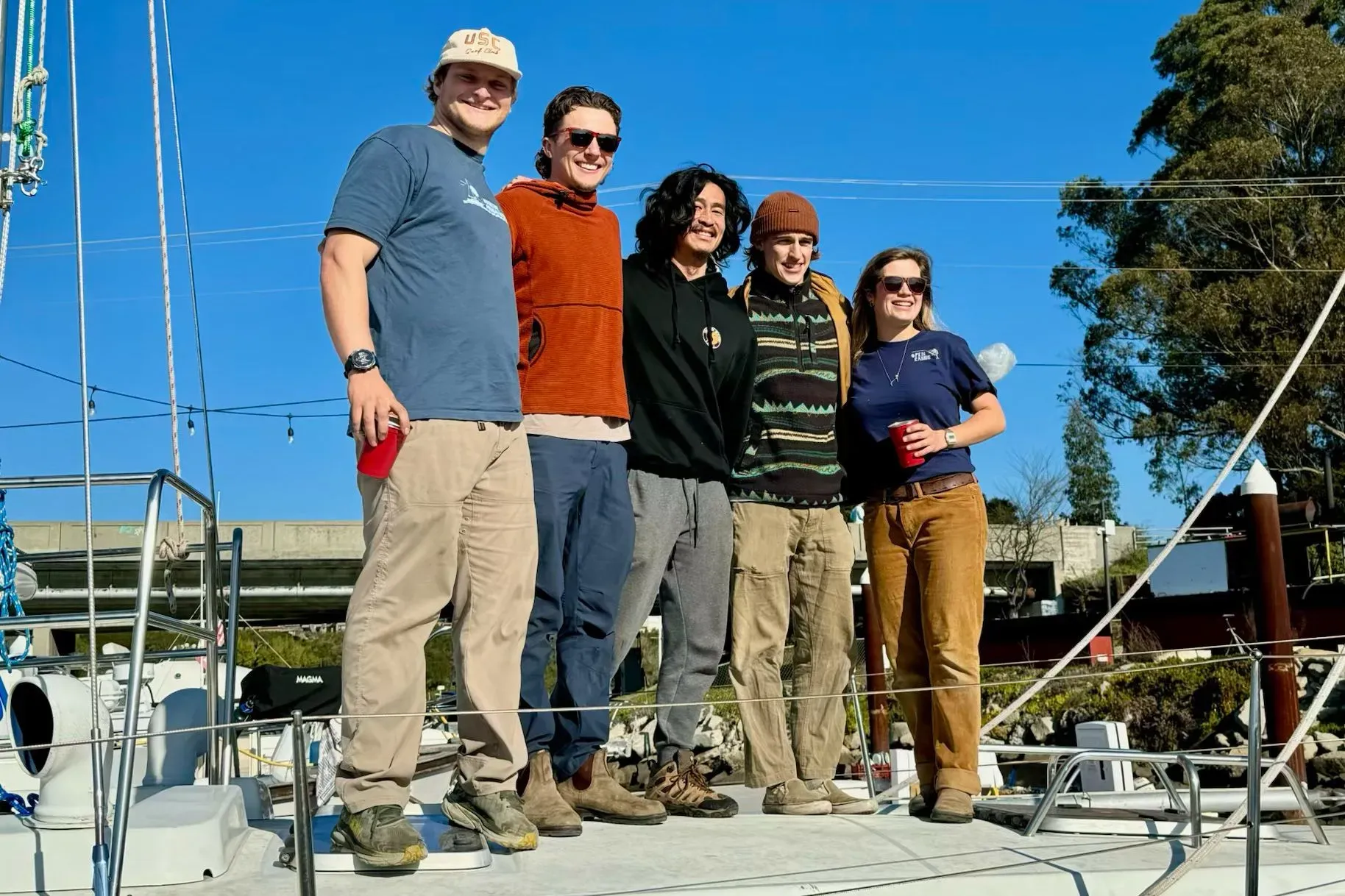 مجموعة من الأصدقاء يقفون معًا على متن قارب شراعي، مبتسمين تحت سماء زرقاء، استعدادًا لمغامرة الإبحار في المحيط الهادئ.