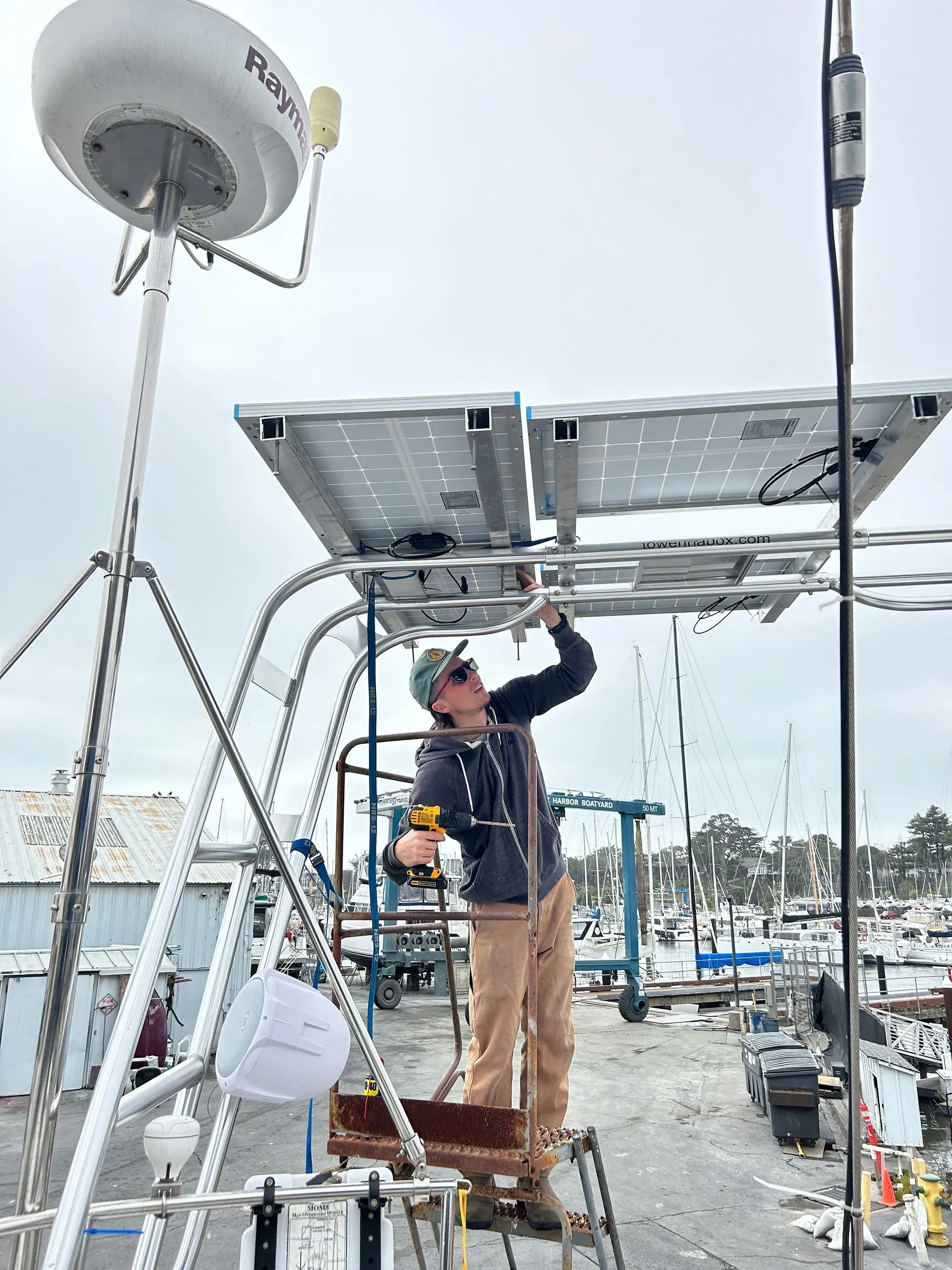 شخص يعمل على تركيب الألواح الشمسية على هيكل قارب شراعي في مرسى، مع قوارب أخرى في الخلفية.