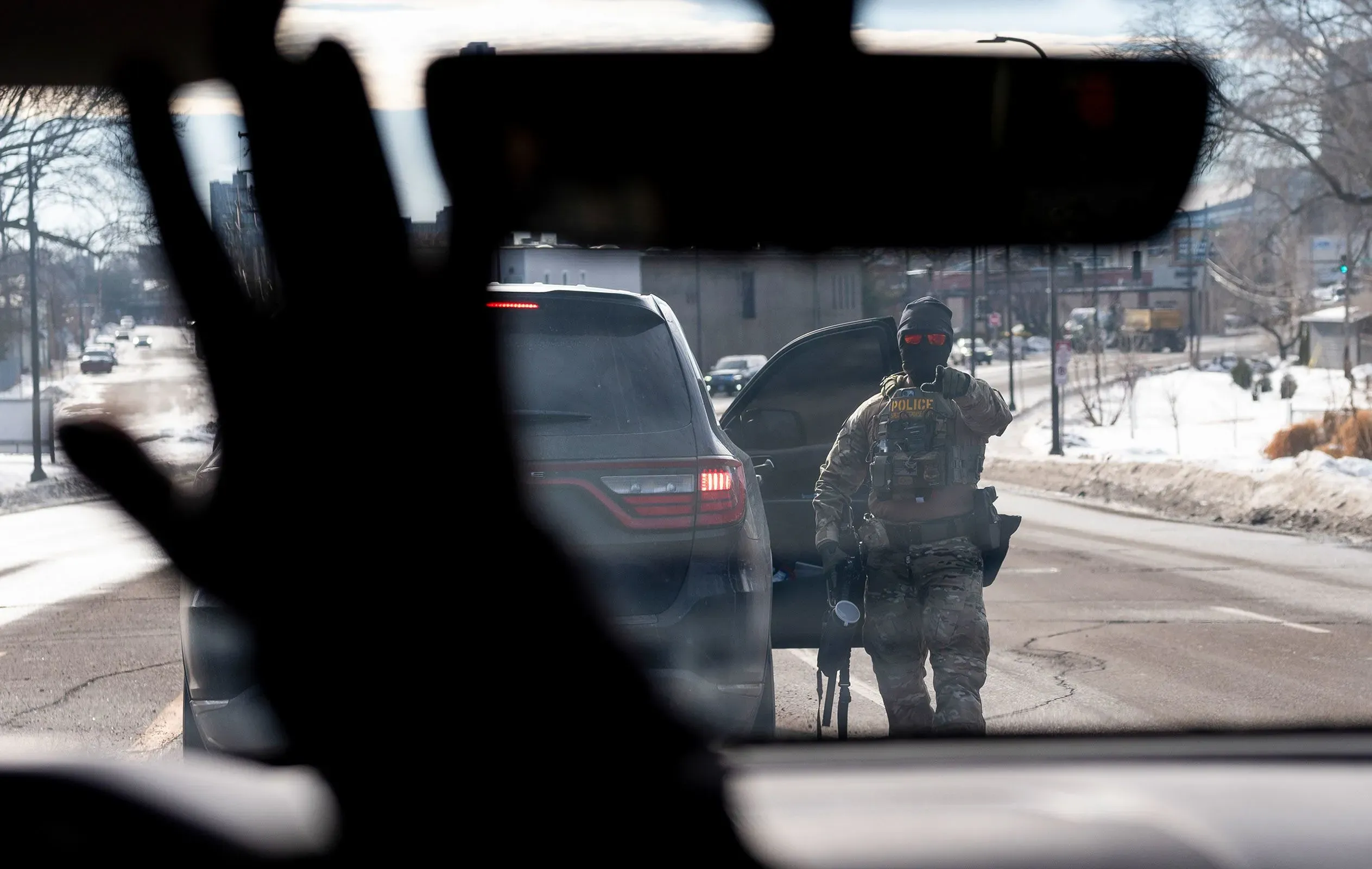 شرطي مسلح يرتدي زيًا عسكريًا يقف بجانب سيارة متوقفة في شارع، مع ظهور يد شخص من داخل السيارة، مما يعكس التوتر في عمليات إنفاذ قوانين الهجرة.