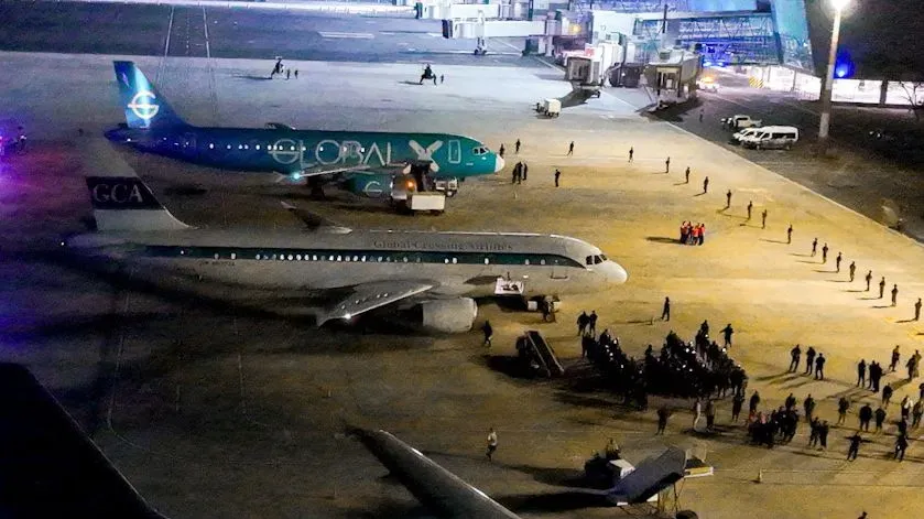 طائرات في مطار، مع وجود مجموعة من الأفراد في انتظار الترحيل إلى السلفادور، مما يعكس التوترات حول سياسة الهجرة الأمريكية.