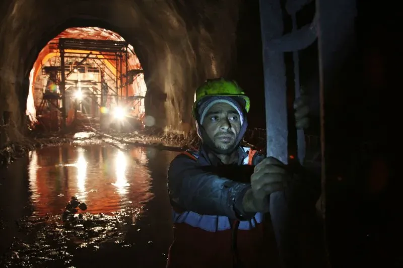 عامل يرتدي خوذة ويعمل داخل نفق قيد الإنشاء في منطقة كشمير، مع إضاءة خلفية توضح تفاصيل البناء.