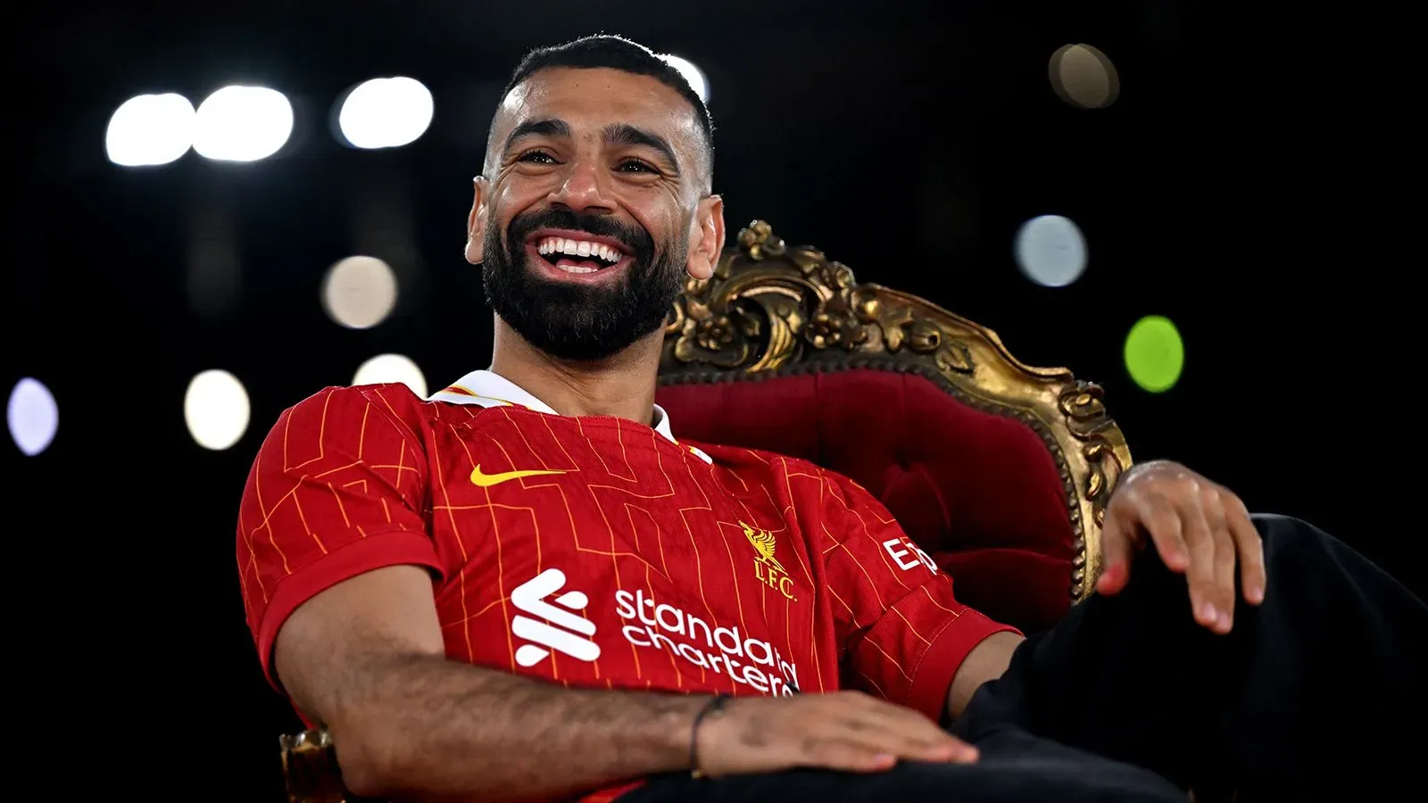 محمد صلاح مبتسم في قميص ليفربول، يجلس على كرسي مميز، مع خلفية مضيئة تعكس أجواء الاحتفال بتوقيع عقده الجديد.