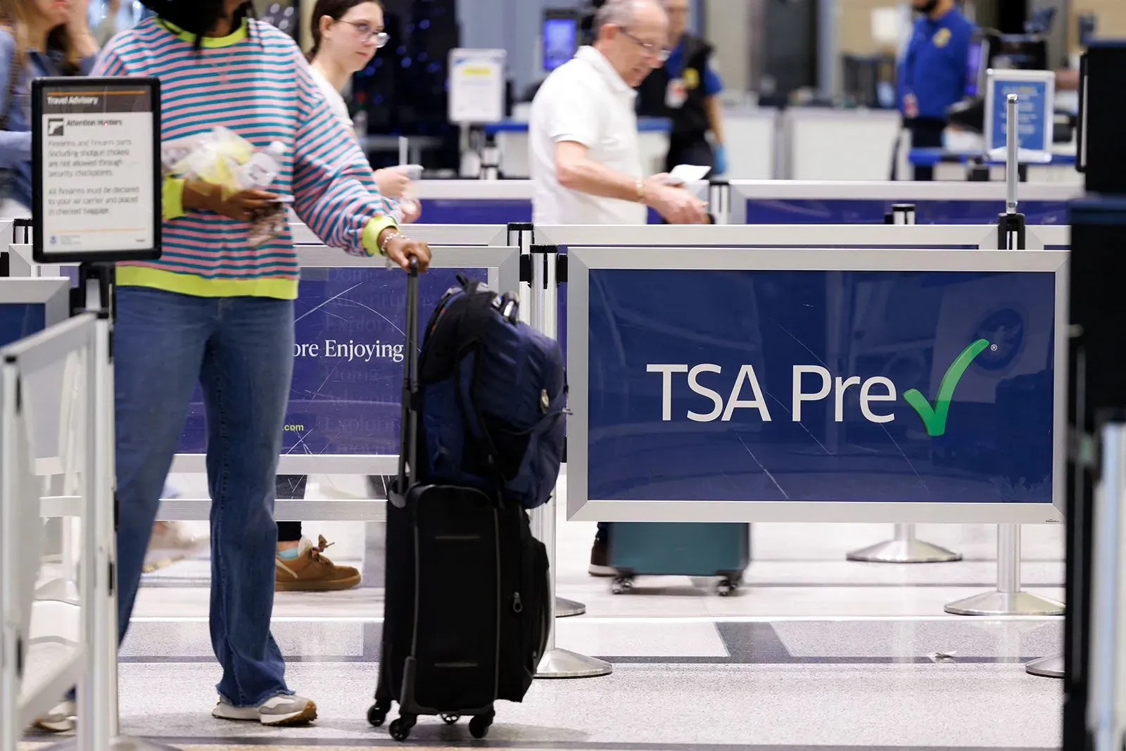 ازدحام في مطار مع وجود لافتة تشير إلى "TSA Pre" بينما يسير مسافرون يحملون حقائبهم، مما يعكس تأثير الإغلاق الجزئي على السفر.